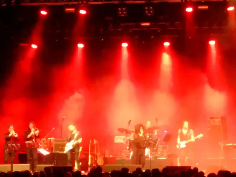 Persönliche Assistenz hilft bei Konzertbesuchen, wo die Location nicht zu 100% barrierefrei ist. So können kleine Stufen überwunden werden. Auf dem Foto zu sehen ein Jazzkonzert mit einer in roten Scheinwerferlicht beleuchteten Bühne. Auf der Bühne stehen rund 10 Musikerinnen und Musiker mit ihren Instrumenten.