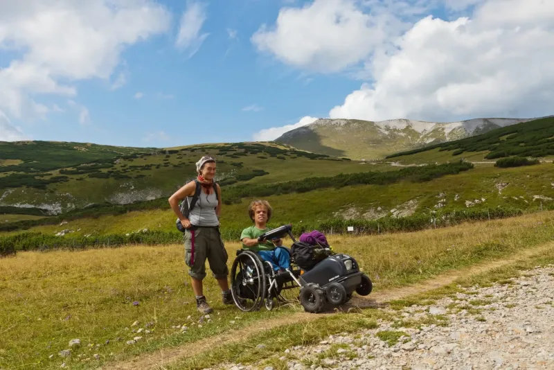Mobilität mit Persönlicher Assistenz. Persönliche Assistenz hilft Rollstuhlfahrer beim Wandern mit einem Swisstrac Zuggerät. Rollstuhlfahrer und Persönliche Assistentin stehen auf einer Alm am Schneeberg. Im Hintergrund blauer Himmel mit weißen Wolken sowie ein Hochalm mit Wiese, Latschen und felsigem Gipfel. Der Rollstuhlfahrer nutzt ein Swisstrac Zuggerät.