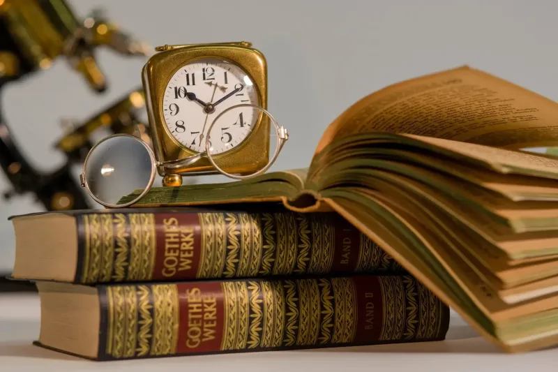 Bücher und Lesestoff neben einer Uhr und Brille eines Persönlichen Assistenten. Während seinen bezahlten Pausen kann der Persönliche Assistent für das Studium lernen.